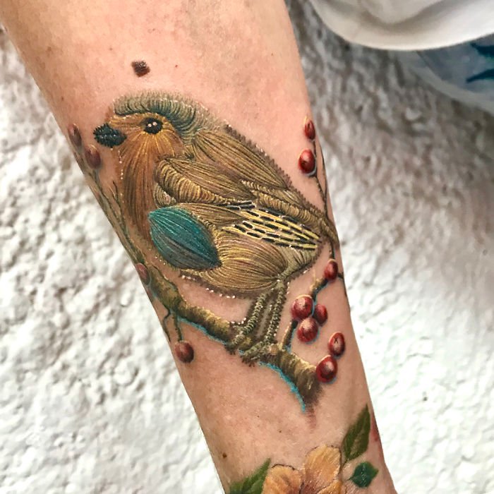 Person showing his arm with a tattoo of brown bird design perched on the branch of a tree with embroidery effect